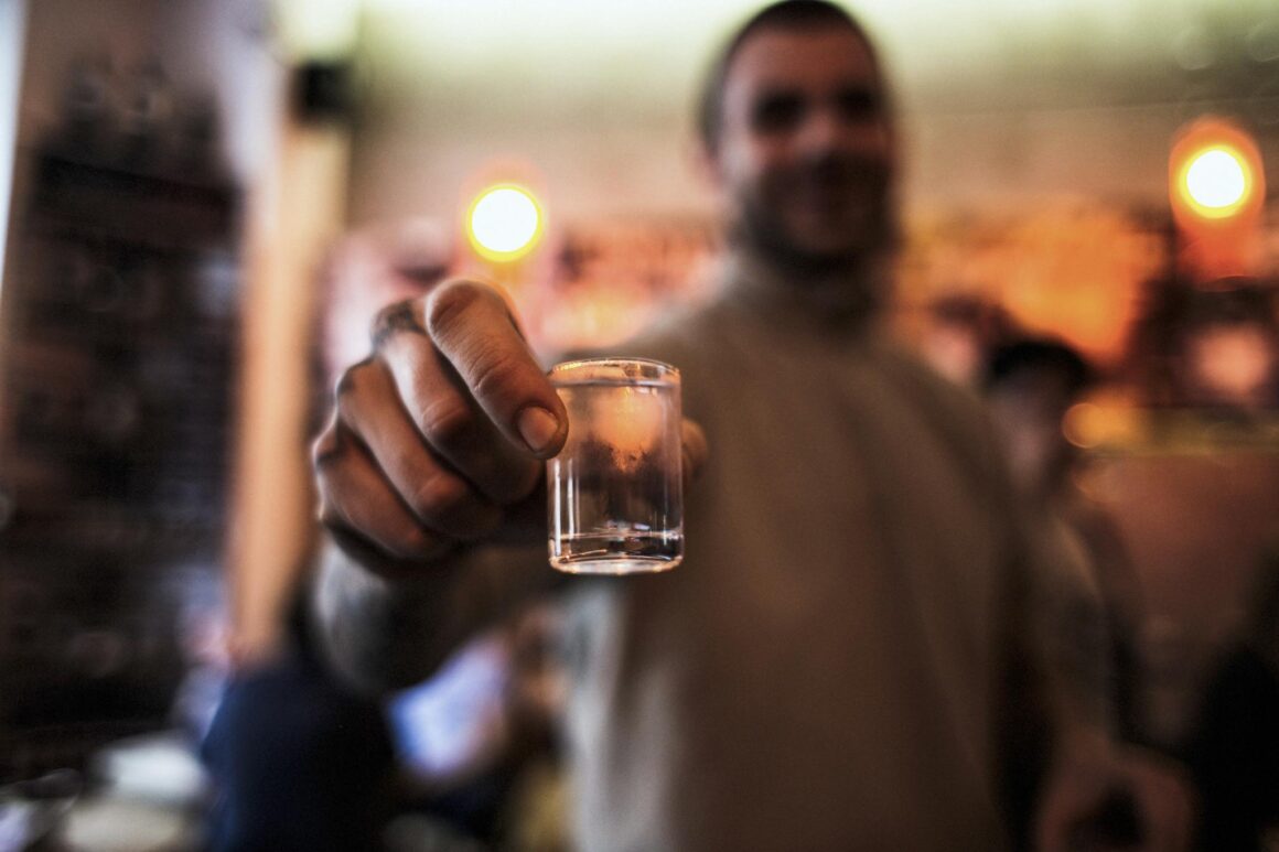 Una persona sostiene un vaso pequeño con una bebida transparente hacia la cámara.